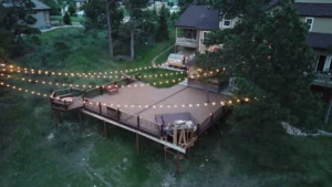 A large elevated wooden deck with string lights is surrounded by trees and grass, in the backyard of a house. The deck has seating areas and a hot tub, creating a cozy, inviting outdoor space in a residential neighborhood.