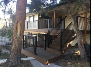 A raised wooden deck with black metal railings and stairs leads to the entrance of a beige house, surrounded by trees and a sloped yard.