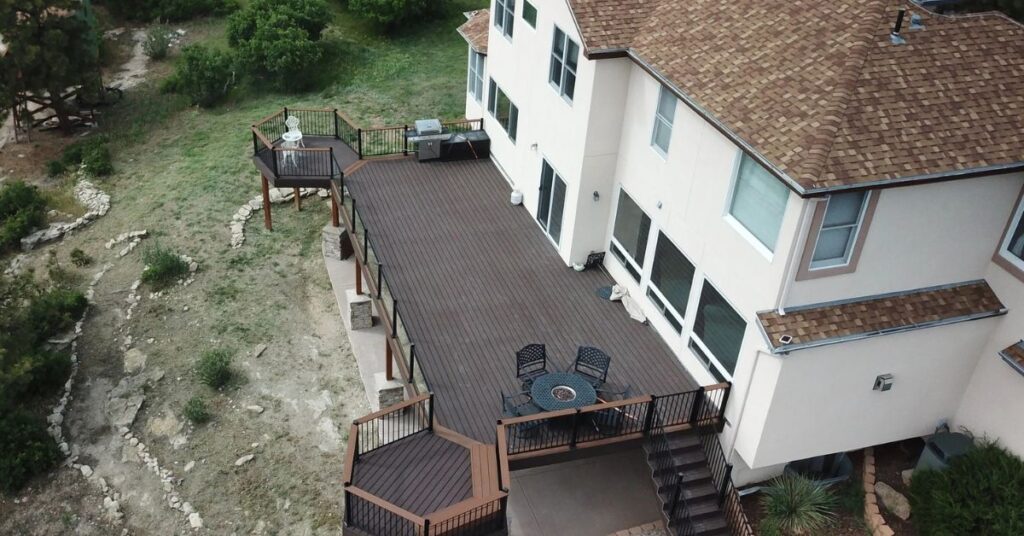 Aerial view of a large house with a spacious wooden deck, featuring patio furniture and multiple staircases, overlooking a grassy yard with trees and shrubs.