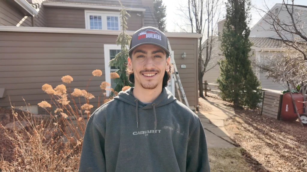 A young man with a mustache wearing a gray Carhartt hoodie and a gray Blue Ridge Builder cap stands smiling in a sunny yard with dried plants and a brown house in the background.