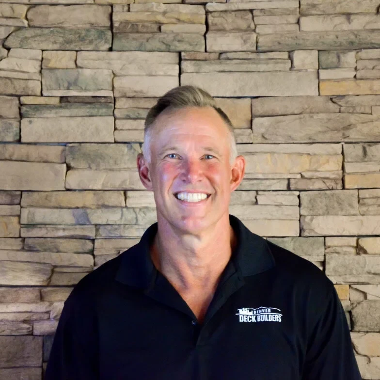 A smiling man with short blonde hair wearing a black collared shirt that reads Deck Builders stands in front of a beige stone wall.