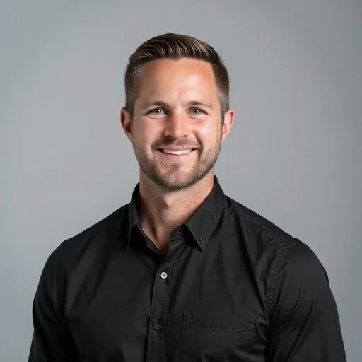 A man with short brown hair and a trimmed beard smiles at the camera while wearing a black button-up shirt against a plain light gray background.